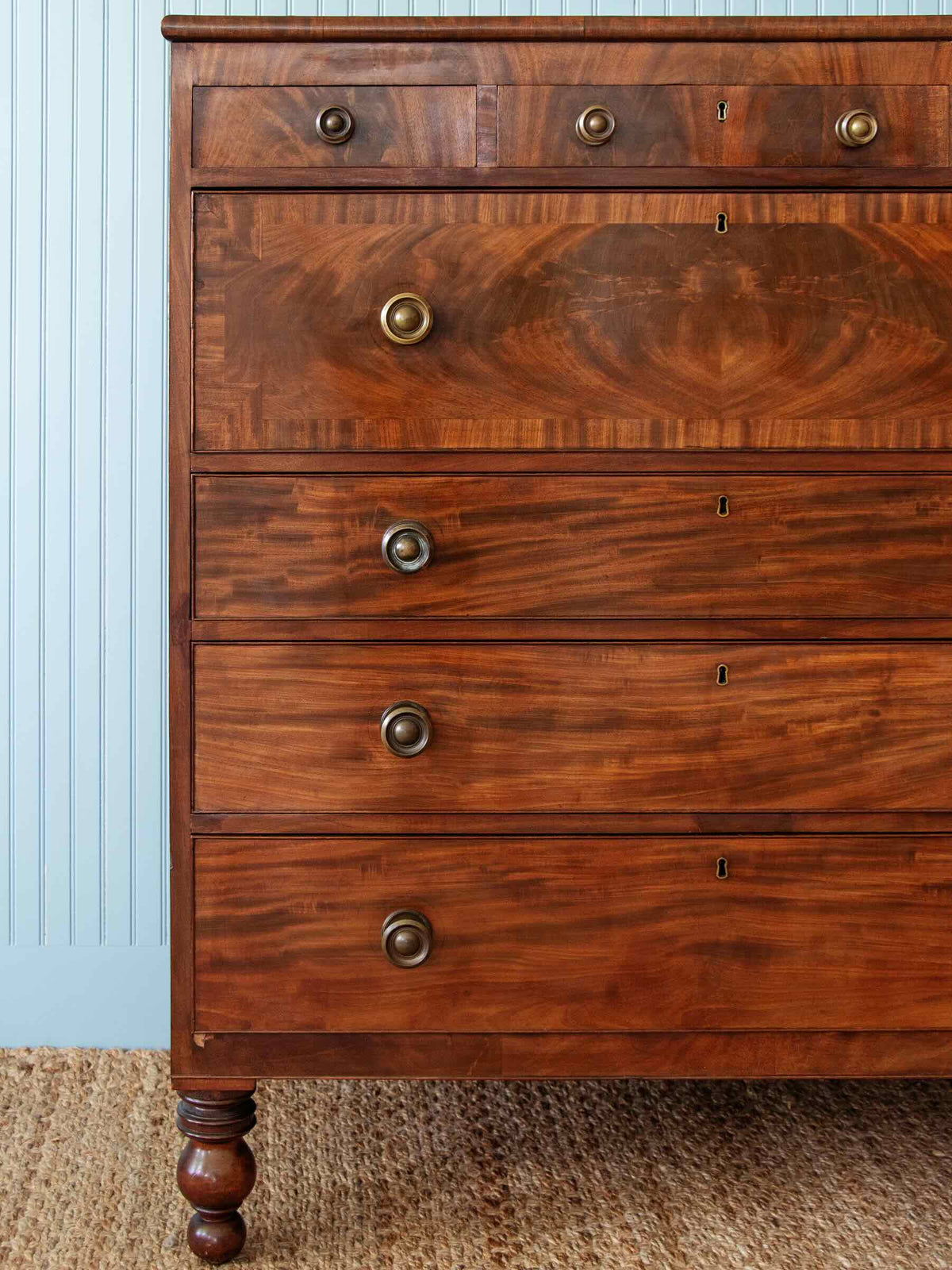 A close-up of a vintage wooden chest of drawers with five drawers, featuring a rich cherry wood grain finish and brass hardware, set against a light blue paneled wall and woven rug.