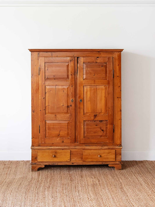 A rustic pine wood double-door cabinet with six paneled doors and two drawers at the bottom, set against a white wall on a woven jute rug.
