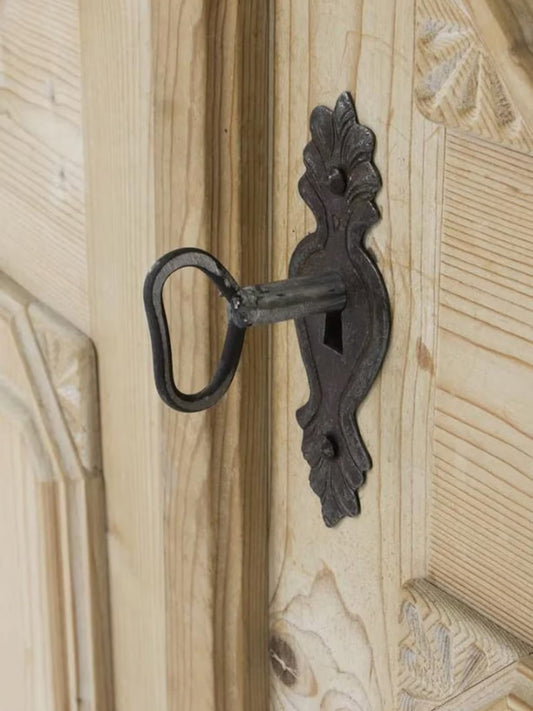 An ornate black metal keyhole with a decorative backplate mounted on a light-colored wooden door.