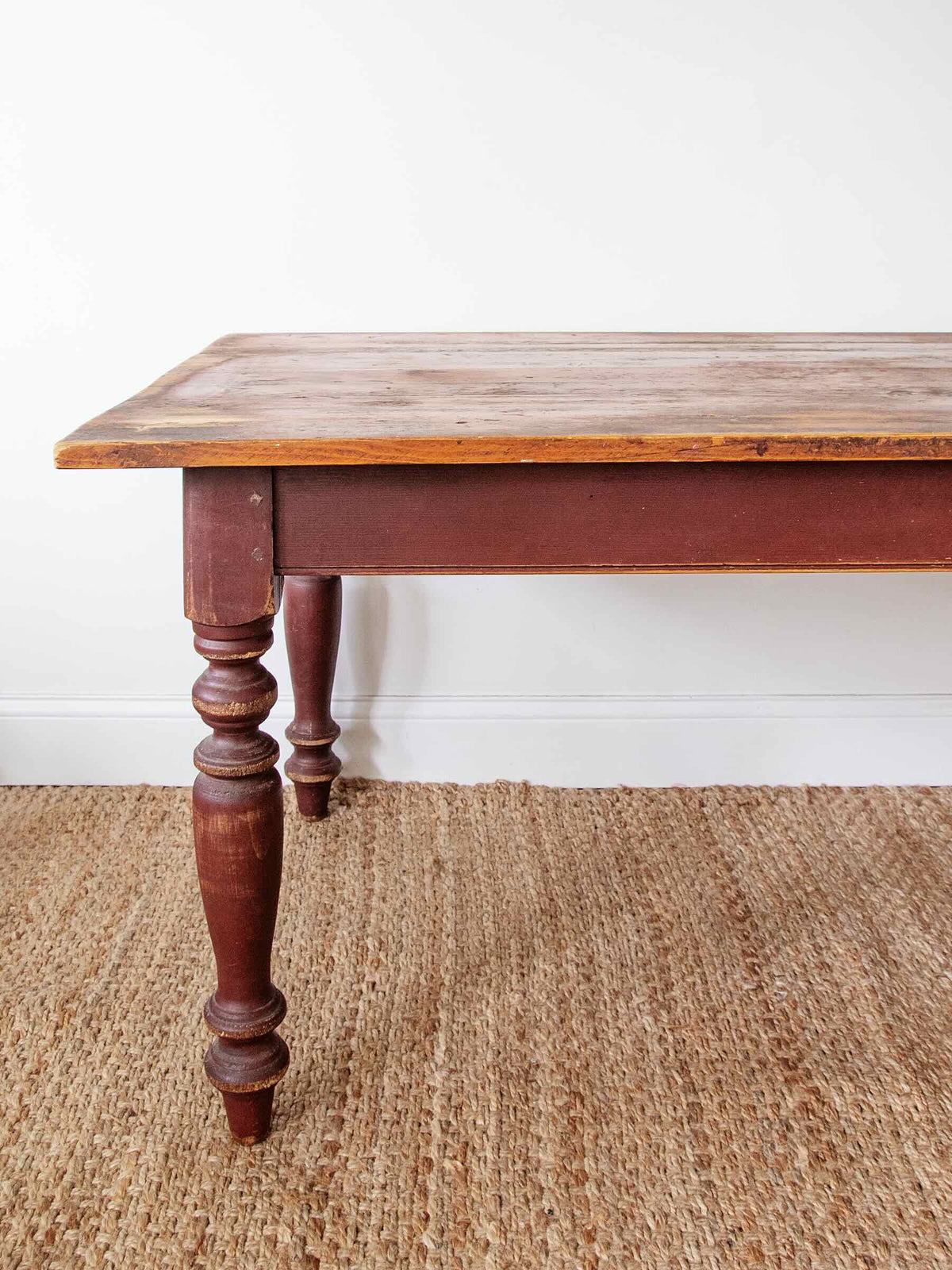 A rustic wooden dining table with a distressed reddish-brown painted frame and turned legs, shown on a woven jute rug against a white wall.