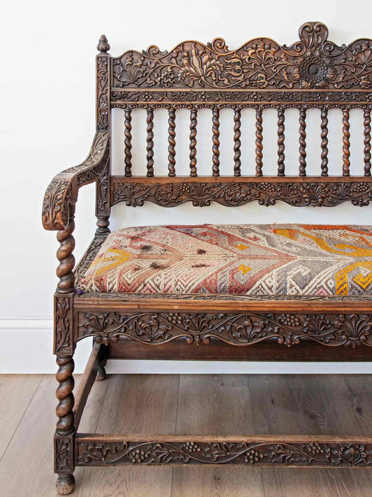 An ornate dark brown carved wooden bench with a patterned beige, red, and yellow upholstered seat cushion, set against a plain white wall and light wood floor.