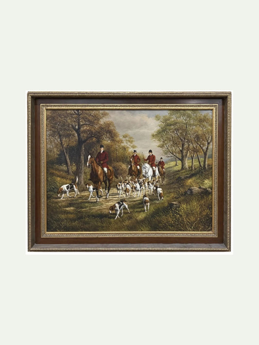A framed landscape painting depicting a fox hunting scene with riders on horseback and hounds in a wooded clearing, shown against a light off-white background.