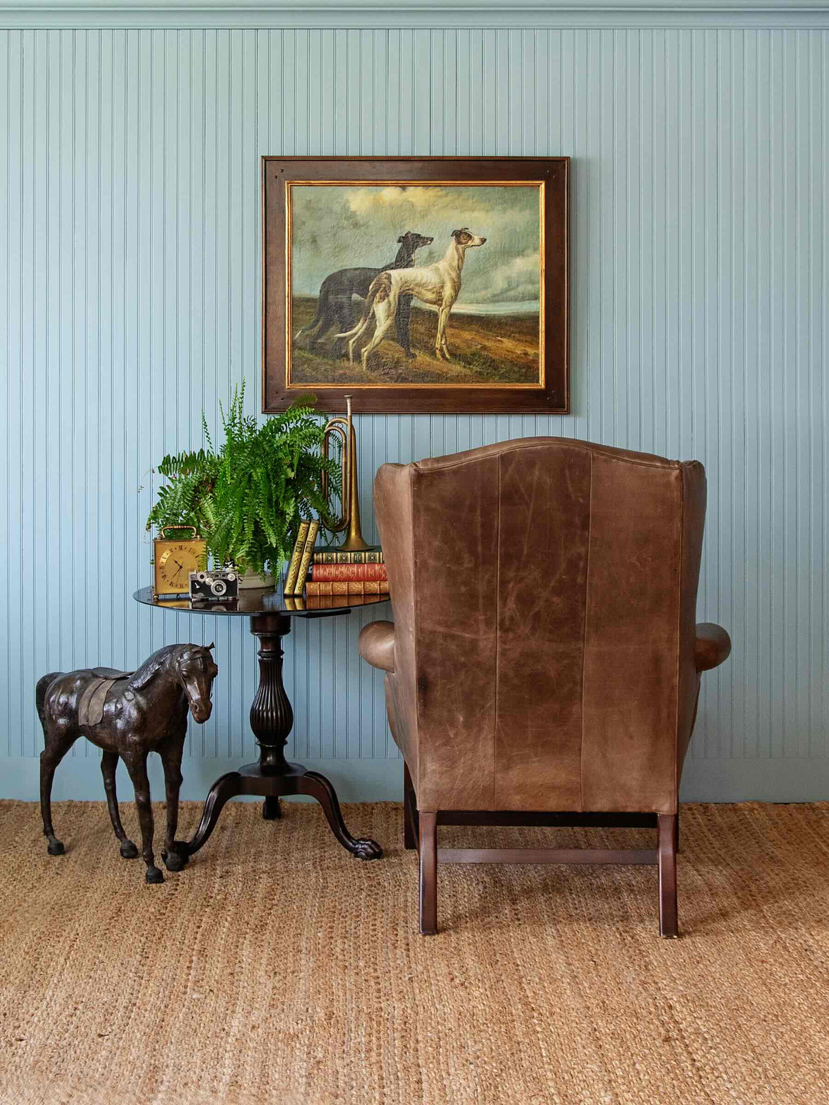 A styled interior scene featuring a brown leather wingback armchair in the foreground, a small dark wood side table with books, a potted fern, and a horse figurine, set against a light blue paneled wall with a framed painting of dogs above.