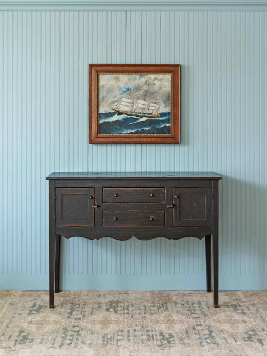 A dark brown wooden console table with three drawers and decorative legs, set against a light blue paneled wall with a framed painting above it.