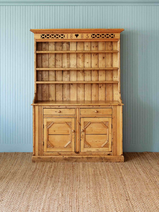 A rustic pine wooden hutch with an open upper shelf section, two drawers, and two cabinet doors with decorative carved panel details, set against a light blue paneled wall and a woven jute rug.