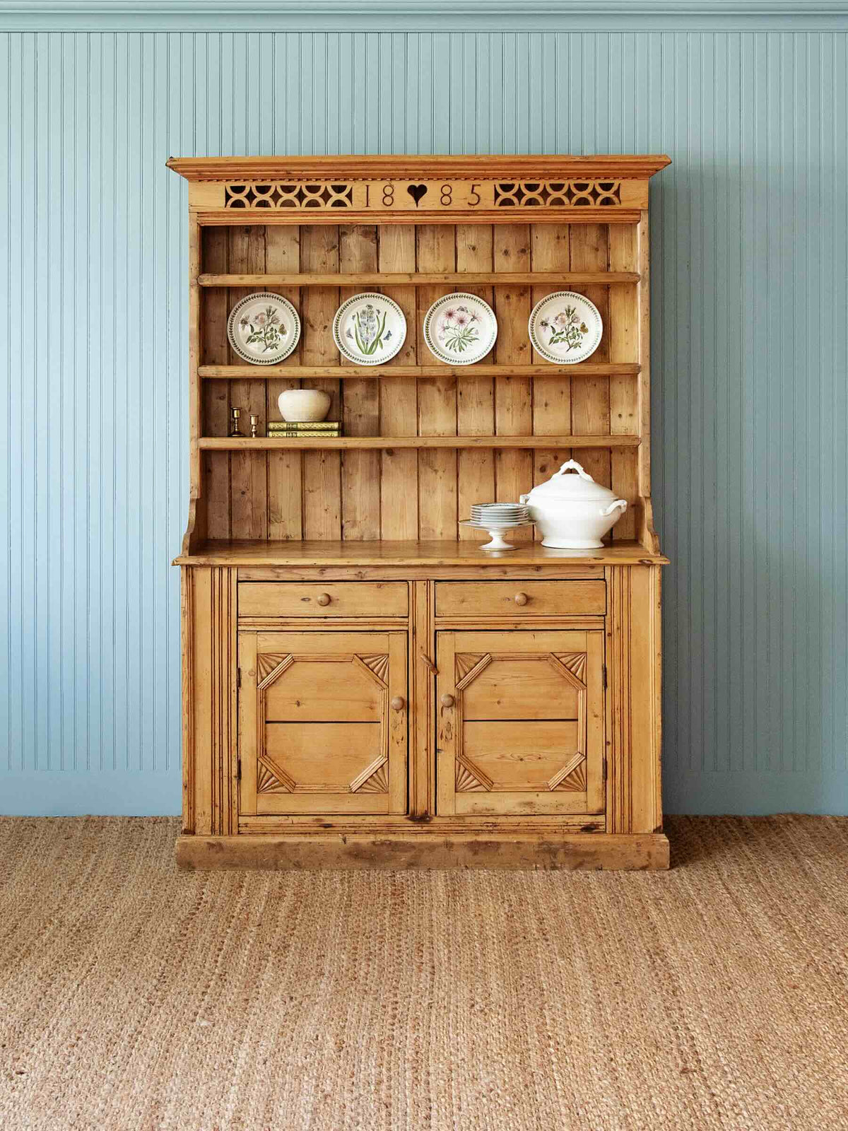 A rustic light brown wooden hutch cabinet with an open upper shelf displaying decorative plates, set against a blue paneled wall and a woven jute rug.