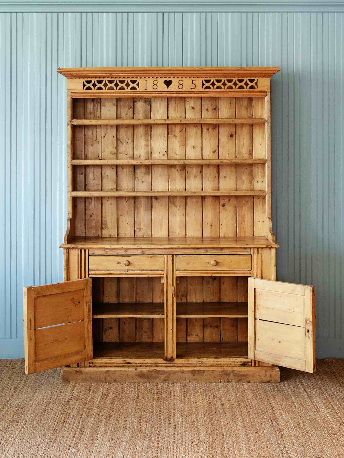 A large rustic wooden hutch cabinet with open doors and multiple shelves, set against a light blue paneled wall and woven jute rug.