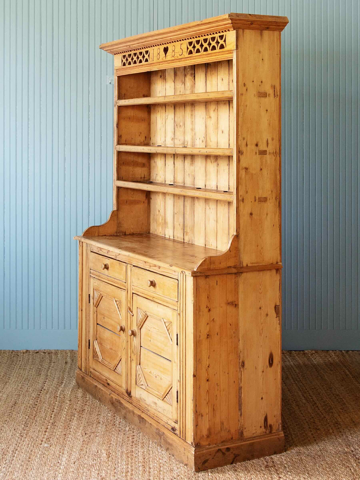 A tall rustic pine hutch with a built-in shelf display, featuring two cabinet doors and drawers on the lower section, set against a blue paneled wall and woven jute rug.