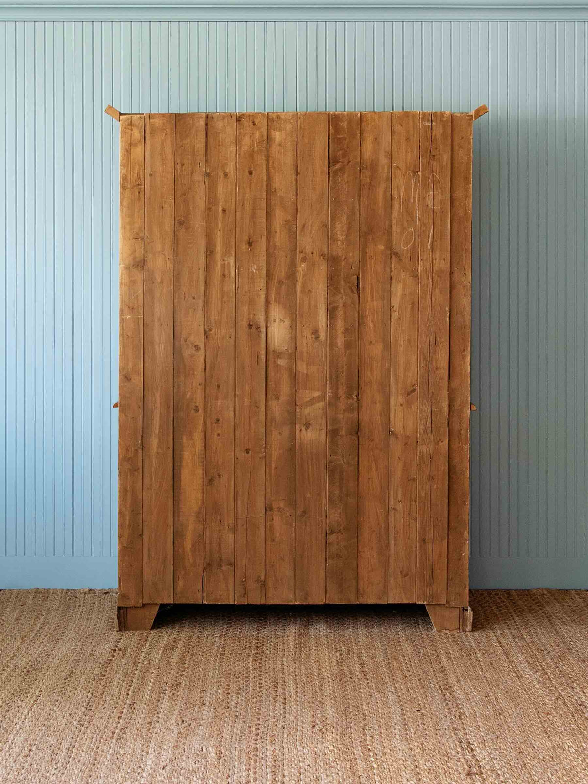 A tall wooden cabinet or wardrobe with two large paneled doors, shown standing on a woven jute rug against a light blue paneled wall.