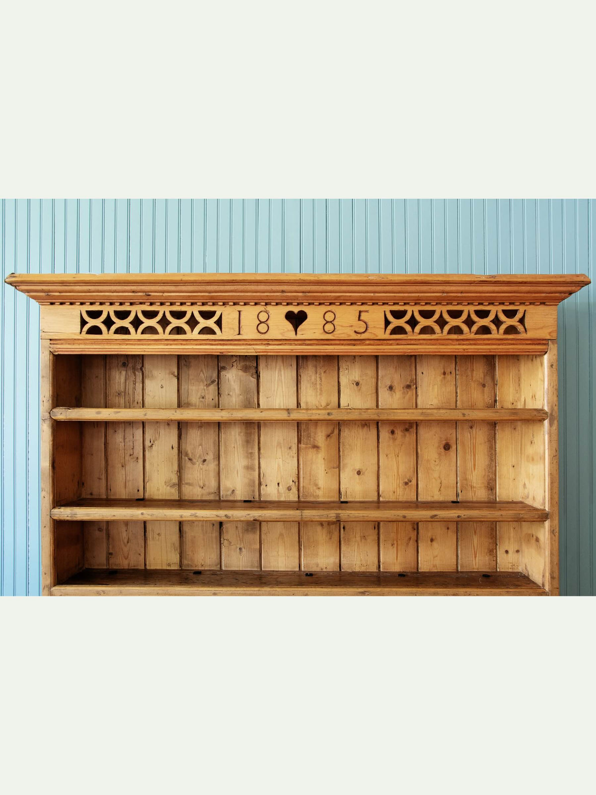 A rustic wooden bookcase with three open shelves, featuring a carved decorative top panel with the numbers 18, a heart, and 85. The bookcase is set against a light blue vertically paneled wall.