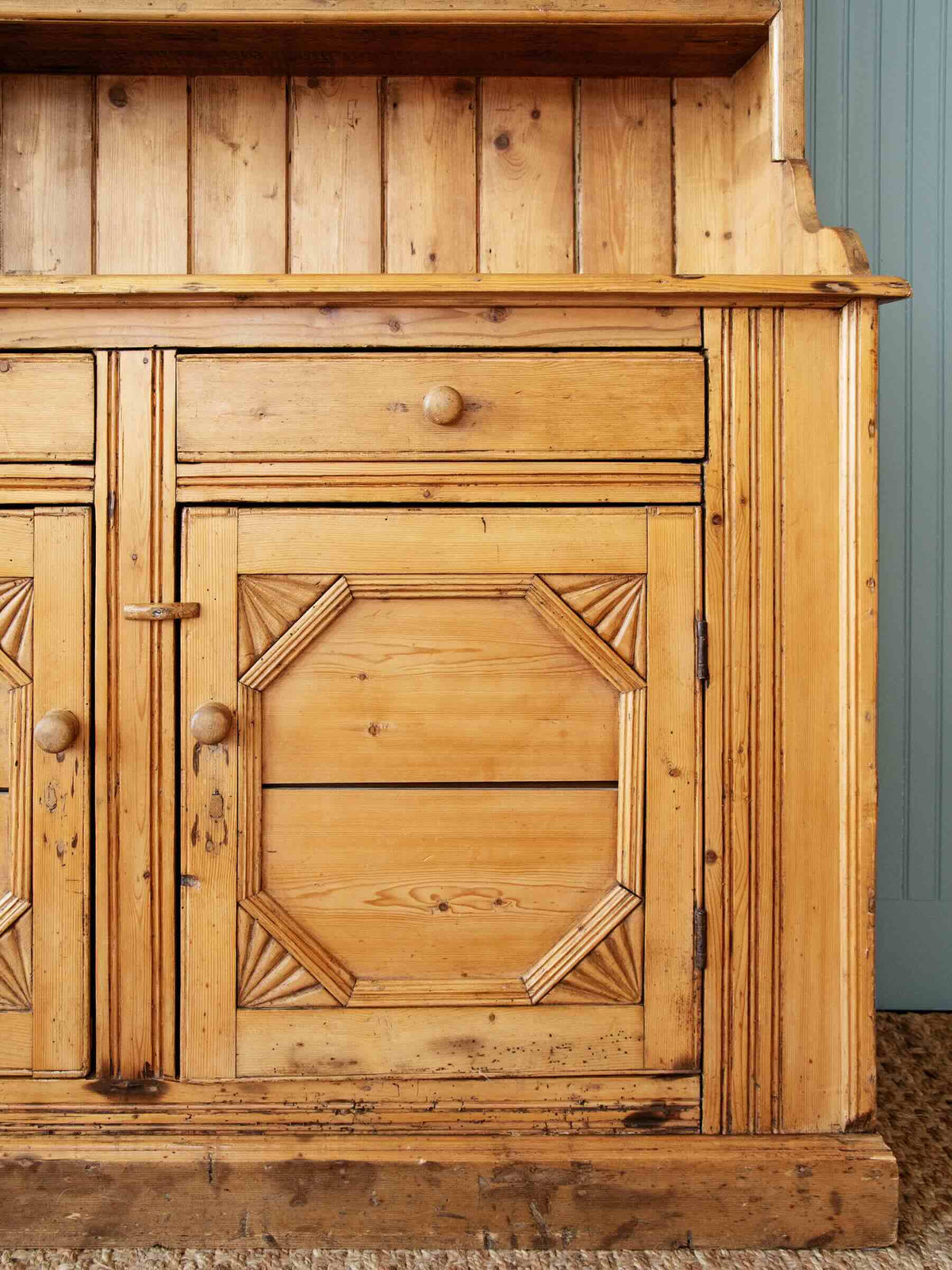 A close-up of a rustic pine wooden cabinet with carved geometric details on the door, a single drawer with a round knob, and a matching side panel, set against a blue paneled wall.