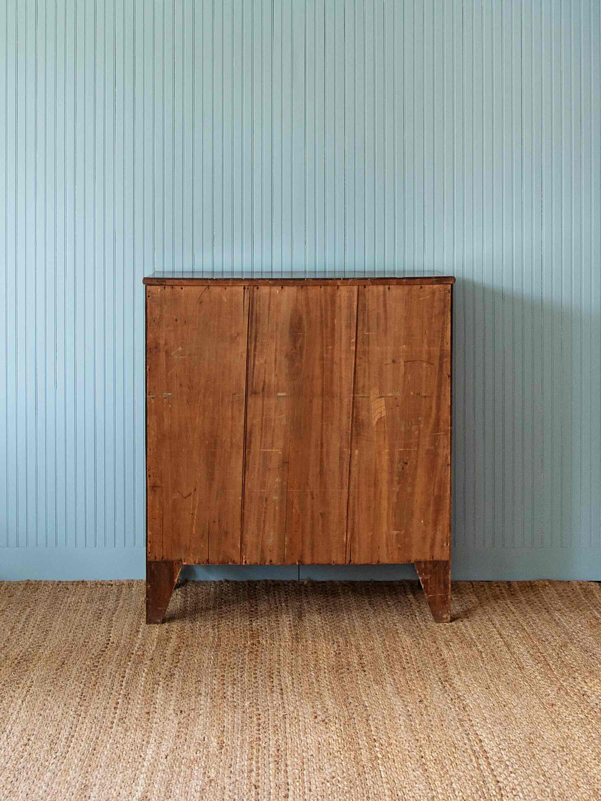 A mid-century modern wooden cabinet with a warm medium-brown finish, featuring vertical panel doors and tapered legs, set against a light blue-green beadboard wall and a natural jute rug.