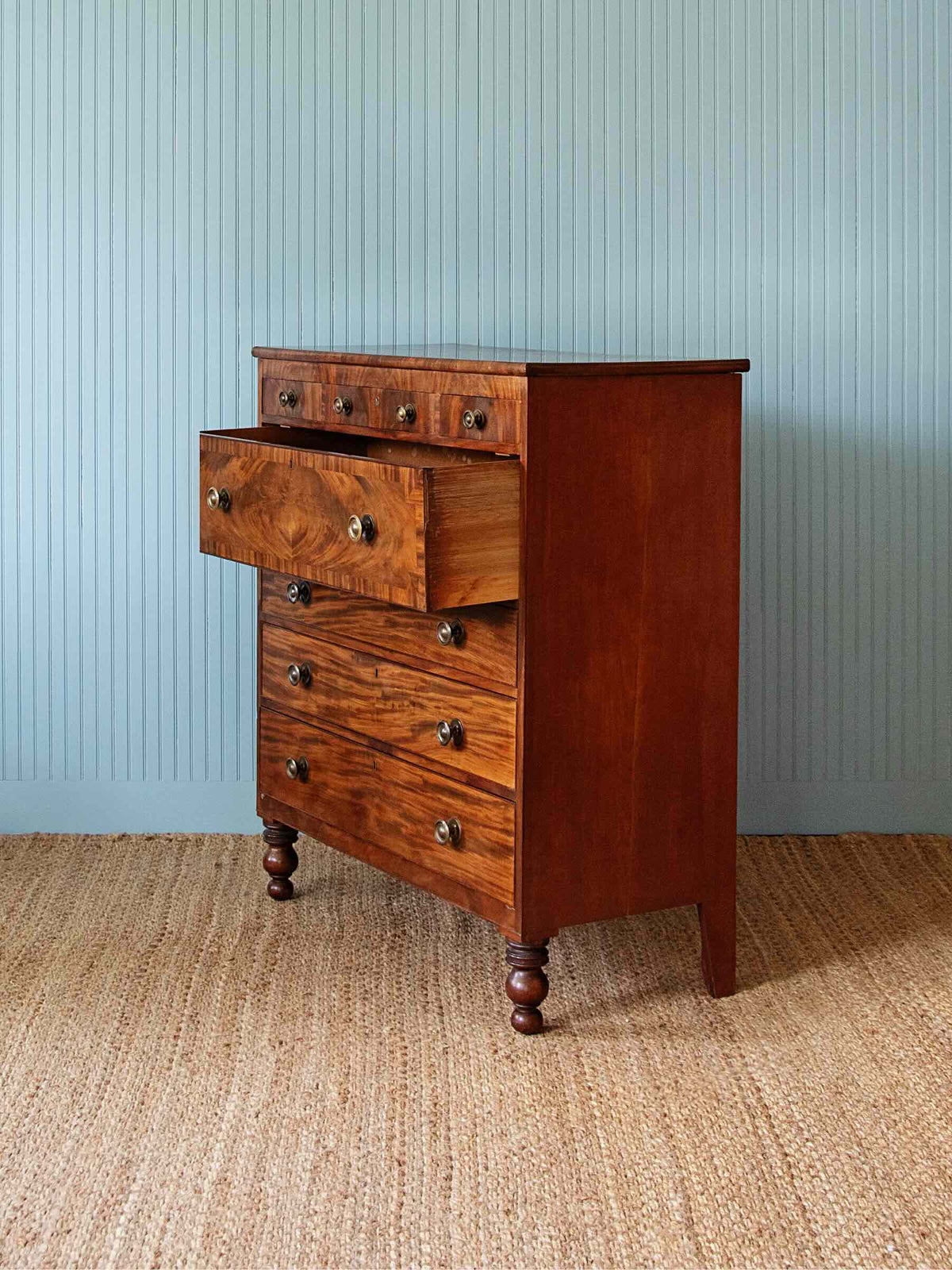 A vintage-style wooden chest of drawers with a rich reddish-brown finish and brass knobs, set against a light blue paneled wall and a woven jute rug.