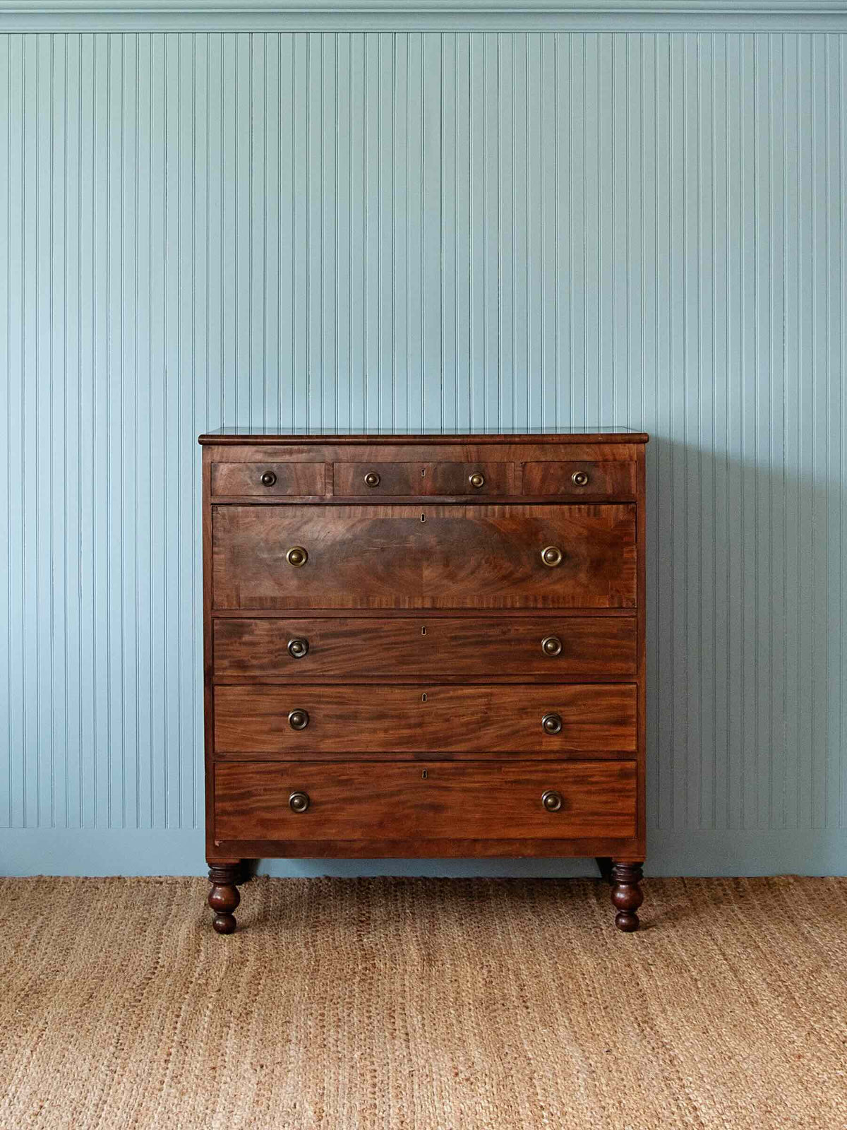 A dark brown wooden five-drawer chest of drawers with brass knobs, set against a light blue paneled wall and a woven jute rug.