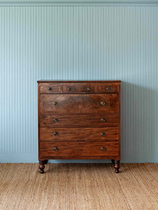 A dark brown wooden five-drawer chest of drawers with brass knobs, set against a light blue paneled wall and a woven jute rug.