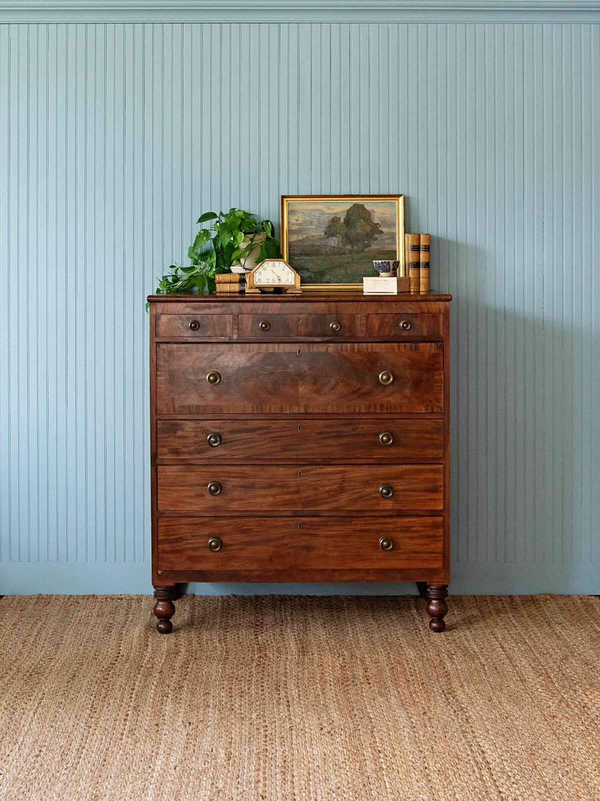 A vintage dark brown wooden chest of drawers with five drawers and brass knobs, styled with a plant, picture frame, and books on top, set against a blue-beaded wall and woven jute rug.