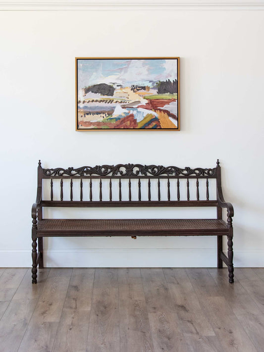A dark brown carved wooden bench with a woven seat, positioned against a white wall below a framed landscape painting on a light wood floor.