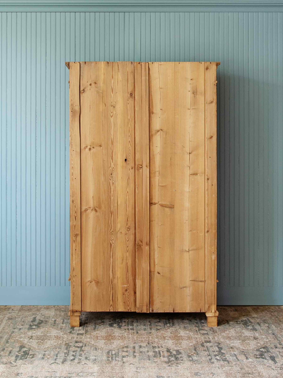 A tall, rustic wooden cabinet with two doors and visible natural wood grain, standing against a light blue paneled wall on a patterned rug.