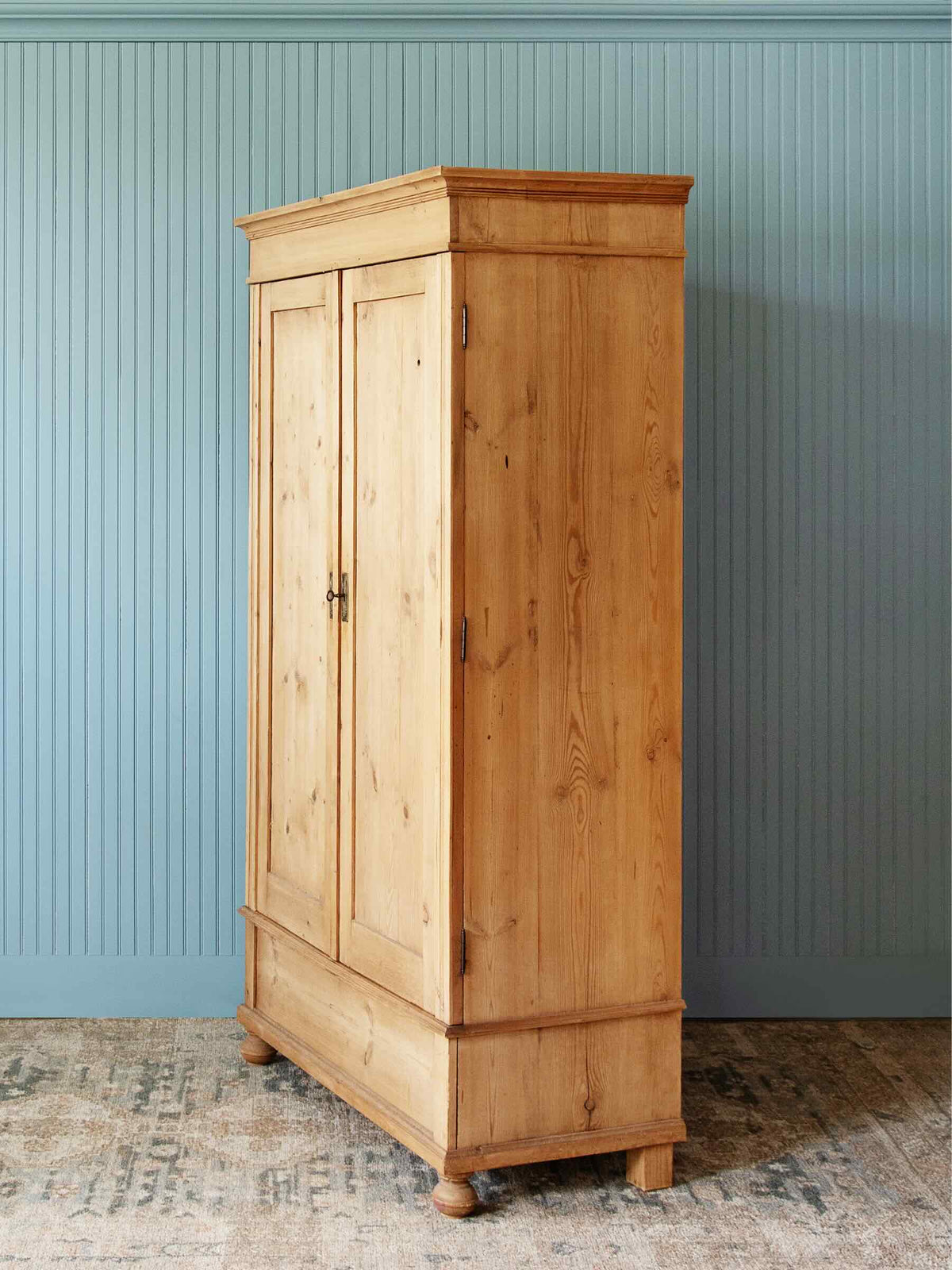A tall, light natural pine double-door wardrobe or armoire cabinet with a decorative top and bottom, standing on a patterned rug against a blue paneled wall.