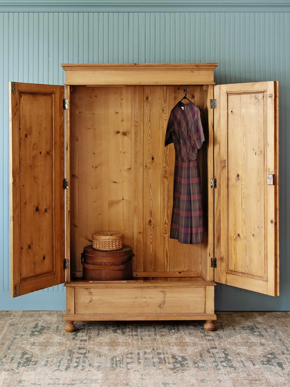 A large rustic pine wooden armoire cabinet with two doors swung open, showing a plaid dress hanging inside, a woven basket, and stacked boxes on the lower shelf, set against a blue paneled wall and patterned rug.