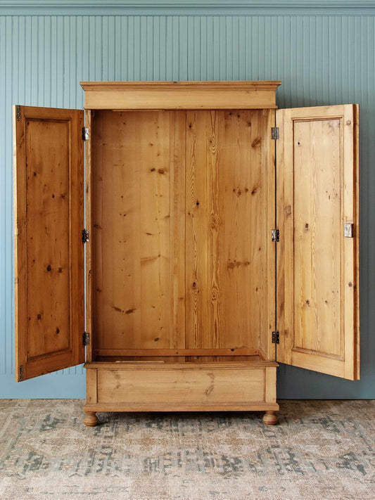 A large natural pine wood armoire cabinet with two doors swung open, showing a tall interior with a lower drawer section, set against a blue paneled wall and patterned rug.