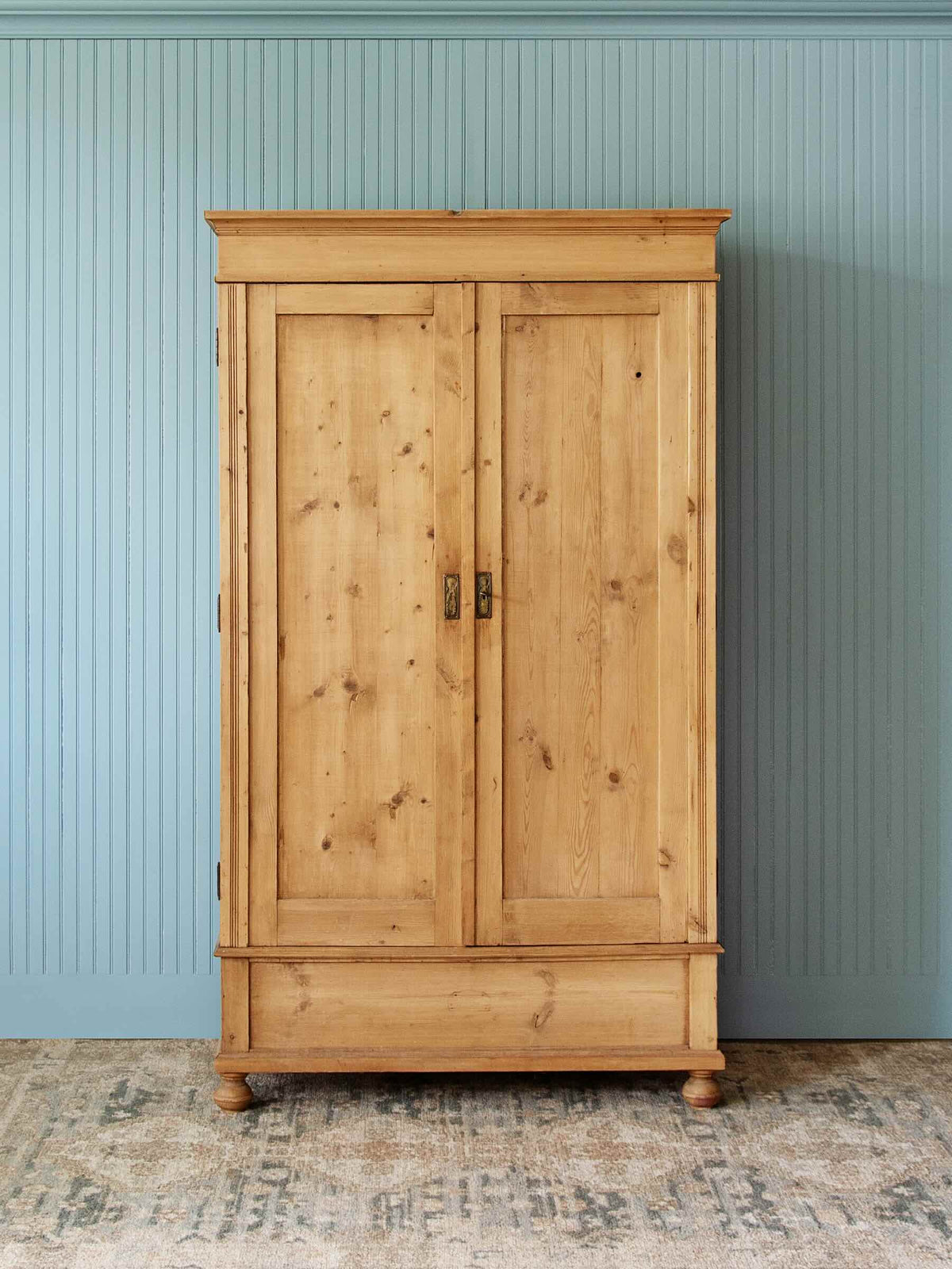 A tall, light natural wood double-door cabinet with dark metal handles, set against a light blue paneled wall and a patterned rug.