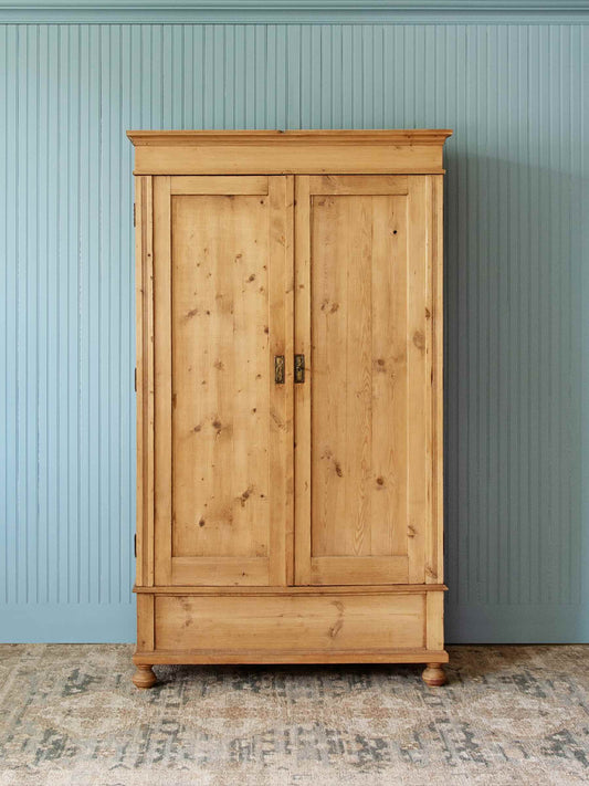 A tall, light natural wood double-door cabinet with dark metal handles, set against a light blue paneled wall and a patterned rug.