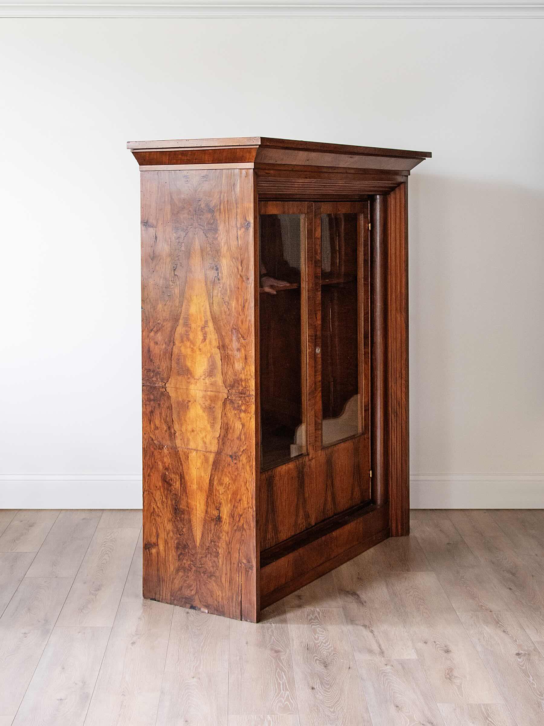 A tall, narrow wooden cabinet with a rich burl wood grain finish and glass-paneled doors, set against a plain white wall and light wood floor.