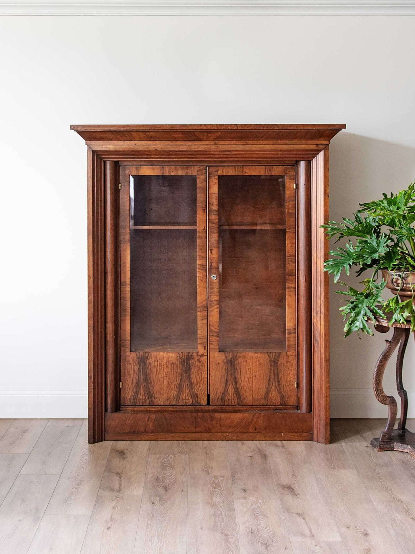A tall, dark brown wooden cabinet with two glass-paneled doors and open shelving, set against a white wall on a light wood floor next to a potted green plant.