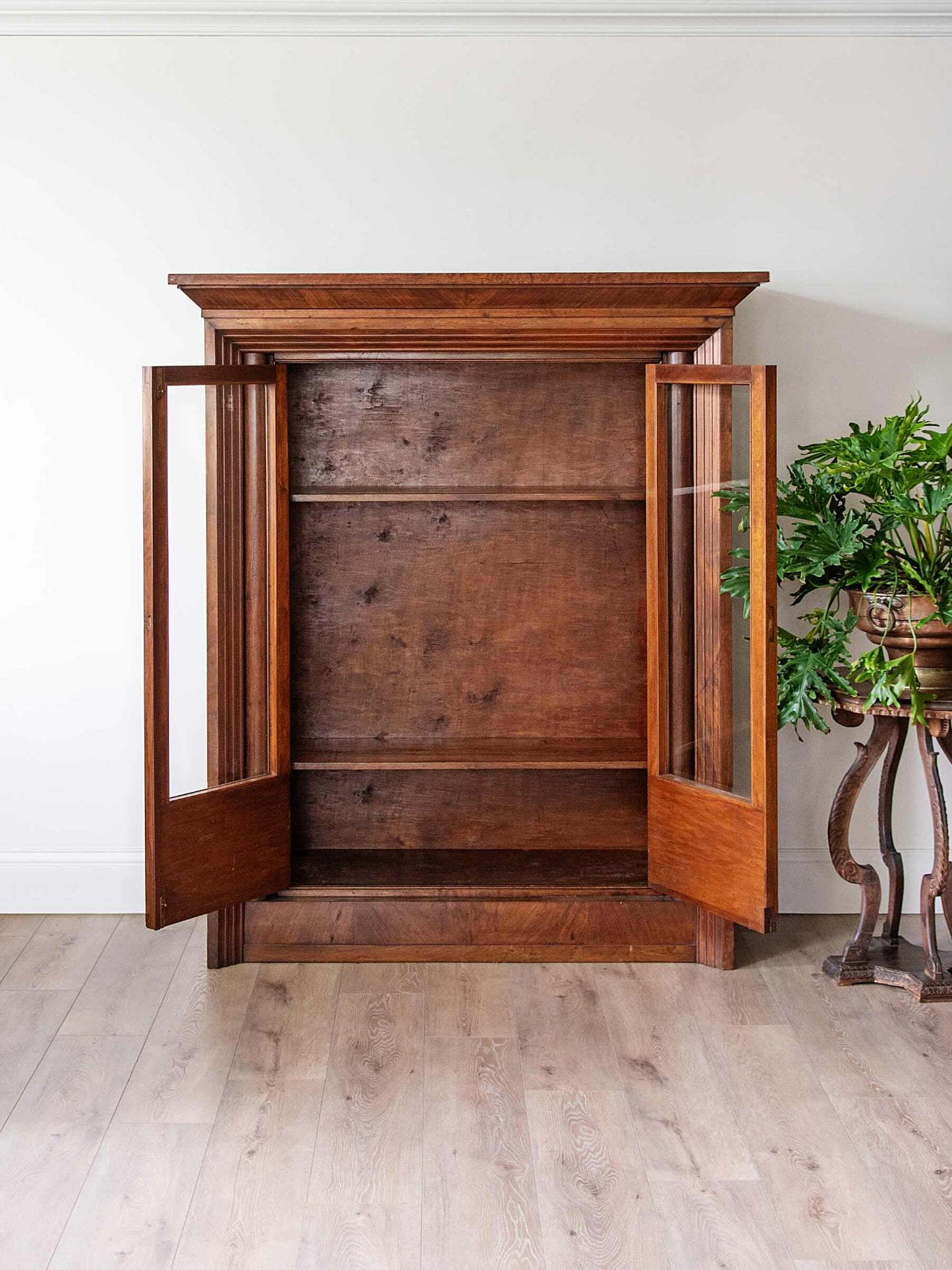 A tall antique wooden cabinet with two open doors and glass panels, set against a white wall on a light wood floor next to a potted green plant.