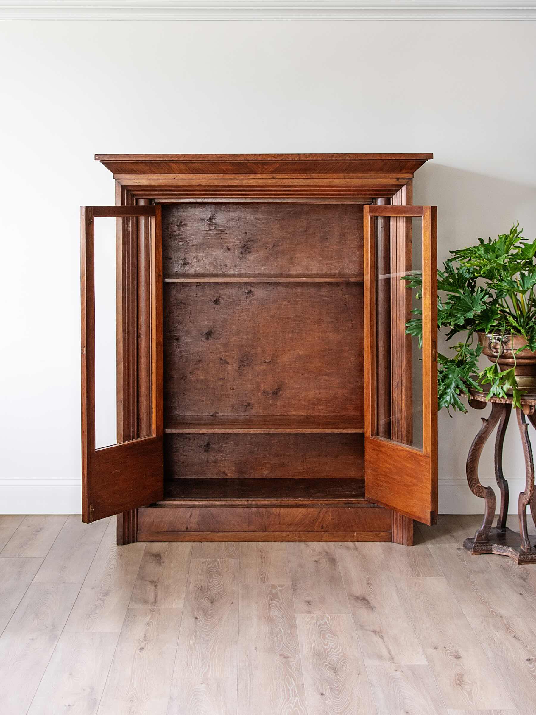 A tall antique wooden cabinet with two open doors and glass panels, set against a white wall on a light wood floor next to a potted green plant.