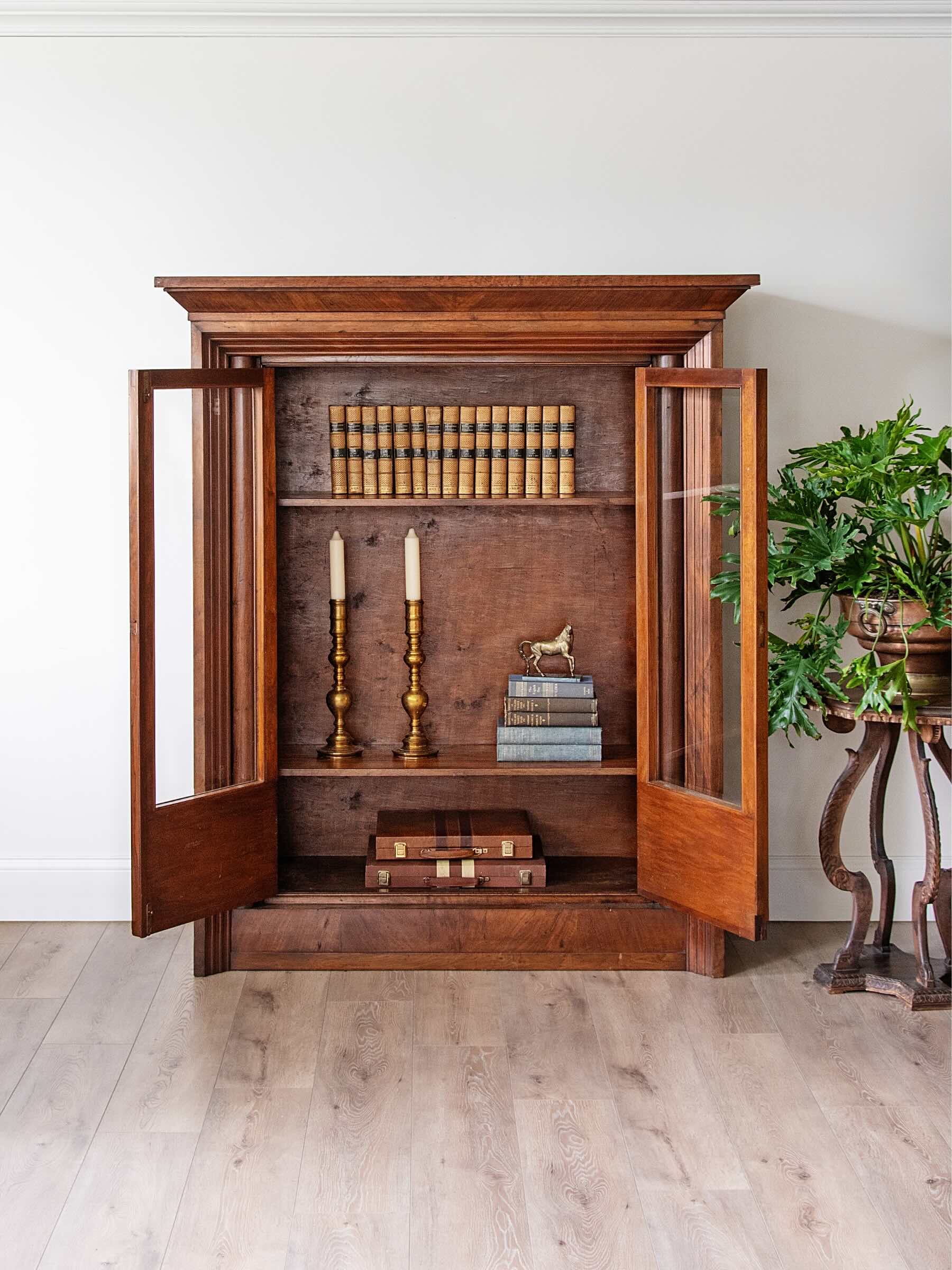 A tall wooden bookcase cabinet with open glass doors, styled with books, candlesticks, and a small horse figurine, set in a room with light wood floors and a potted plant on the right.