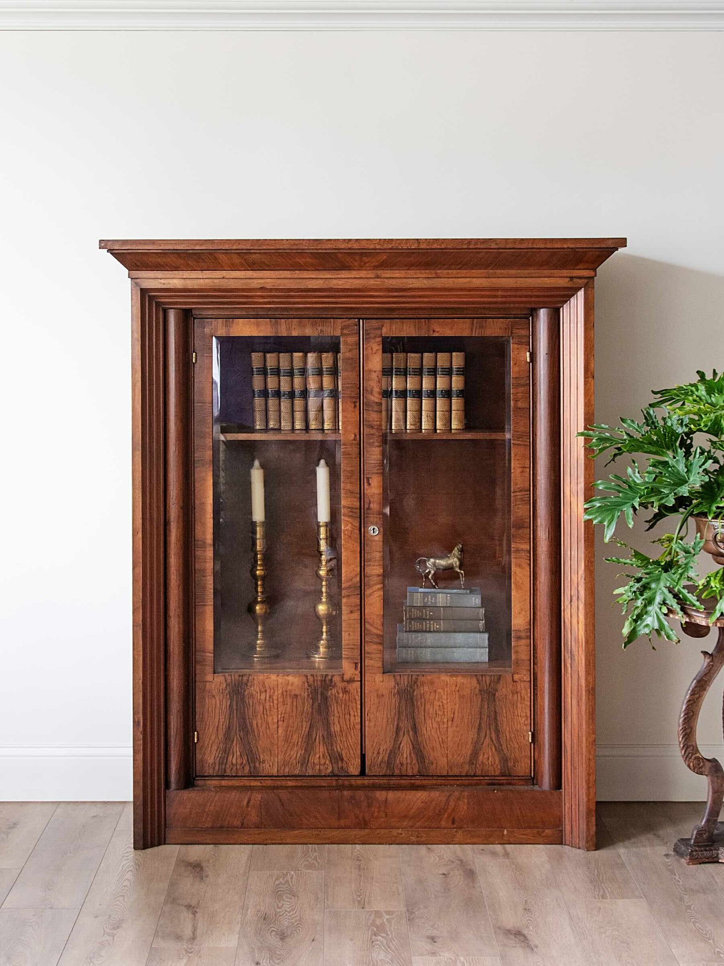 A tall, dark wood bookcase with two glass-paneled doors, styled with books and decorative items, set against a white wall on a light wood floor next to a potted plant.