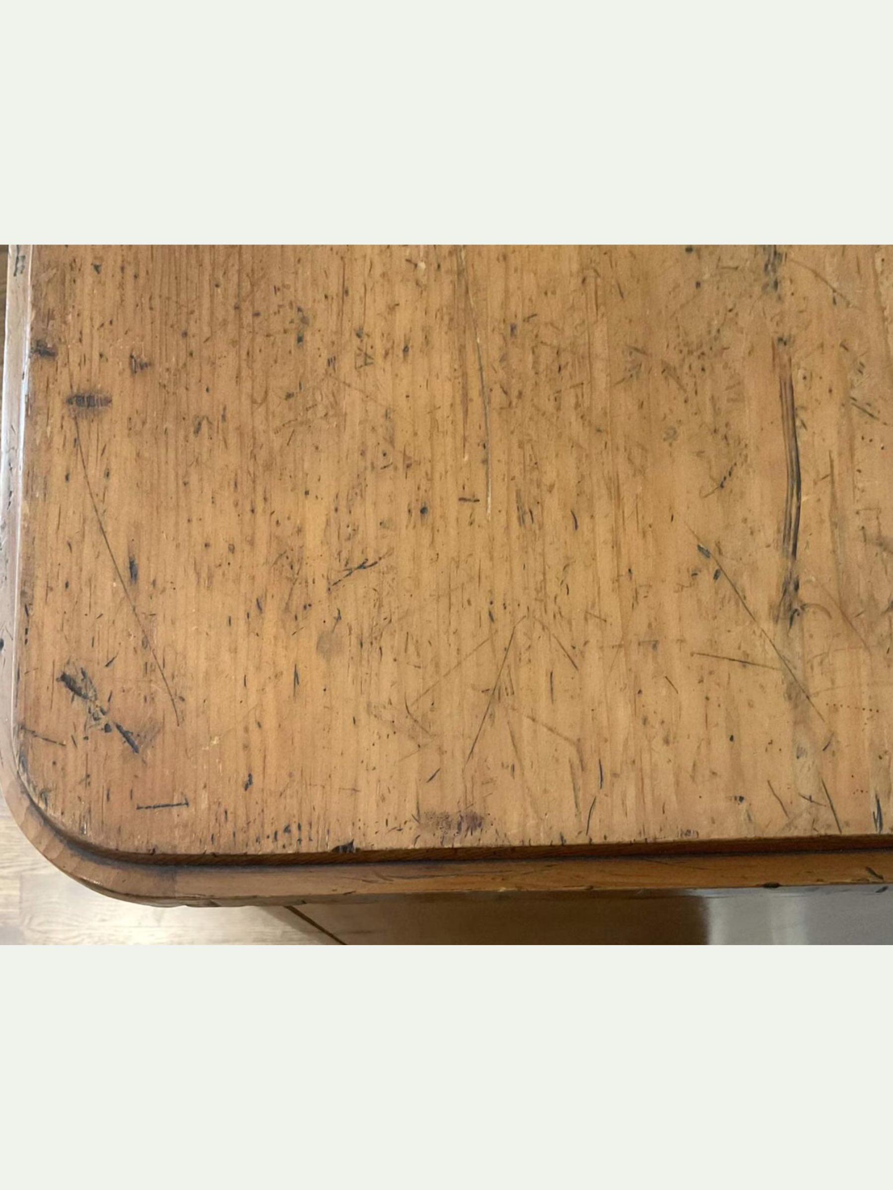 A close-up view of a worn wooden tabletop with visible scratches, scuffs, and dark marks on a light wood frame, set against a plain off-white background.