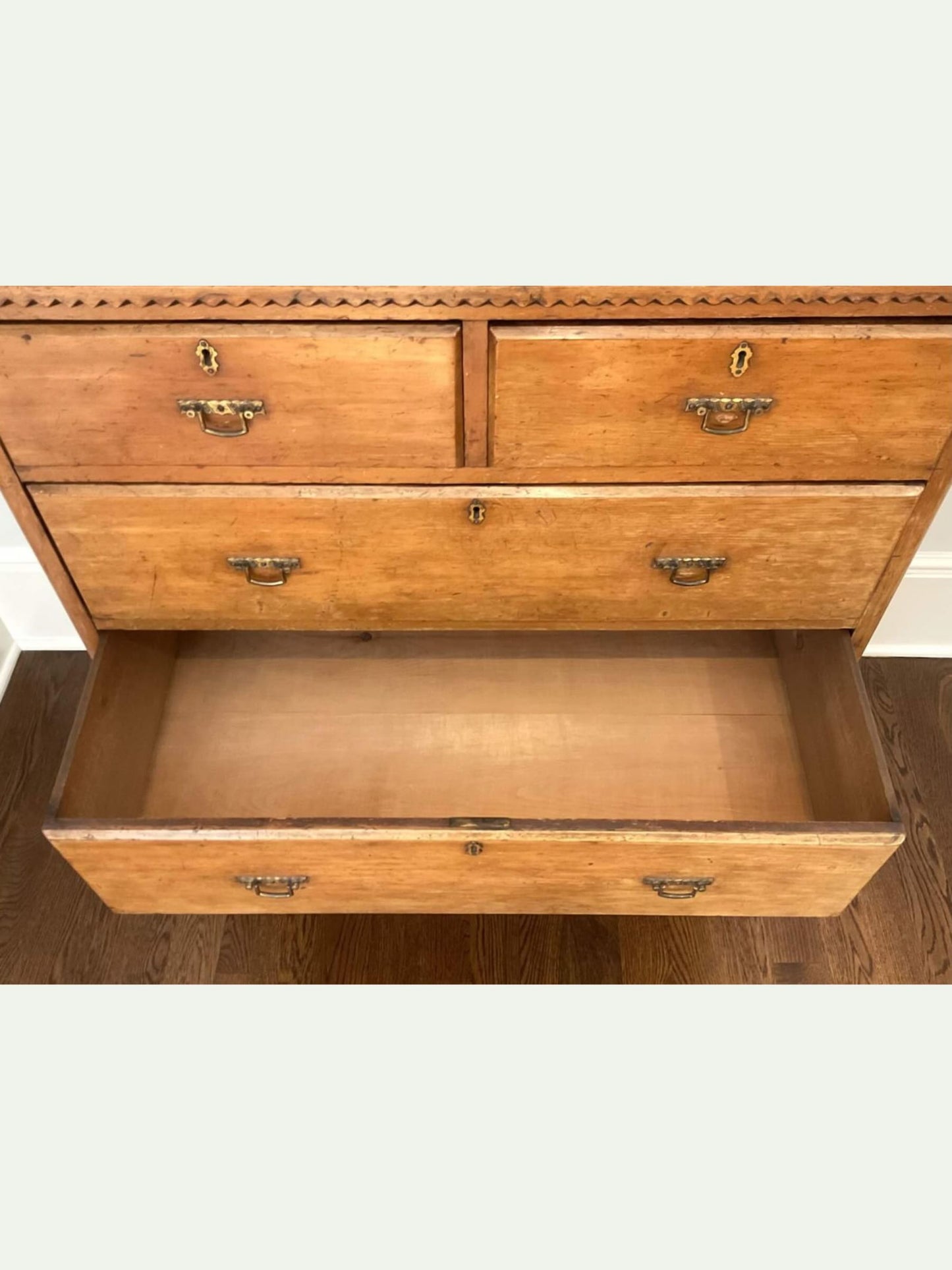 An antique wooden chest of drawers with three drawers, one pulled open, showing its interior. The drawers feature brass handles and a decorative geometric border along the top front, set against a plain light background.