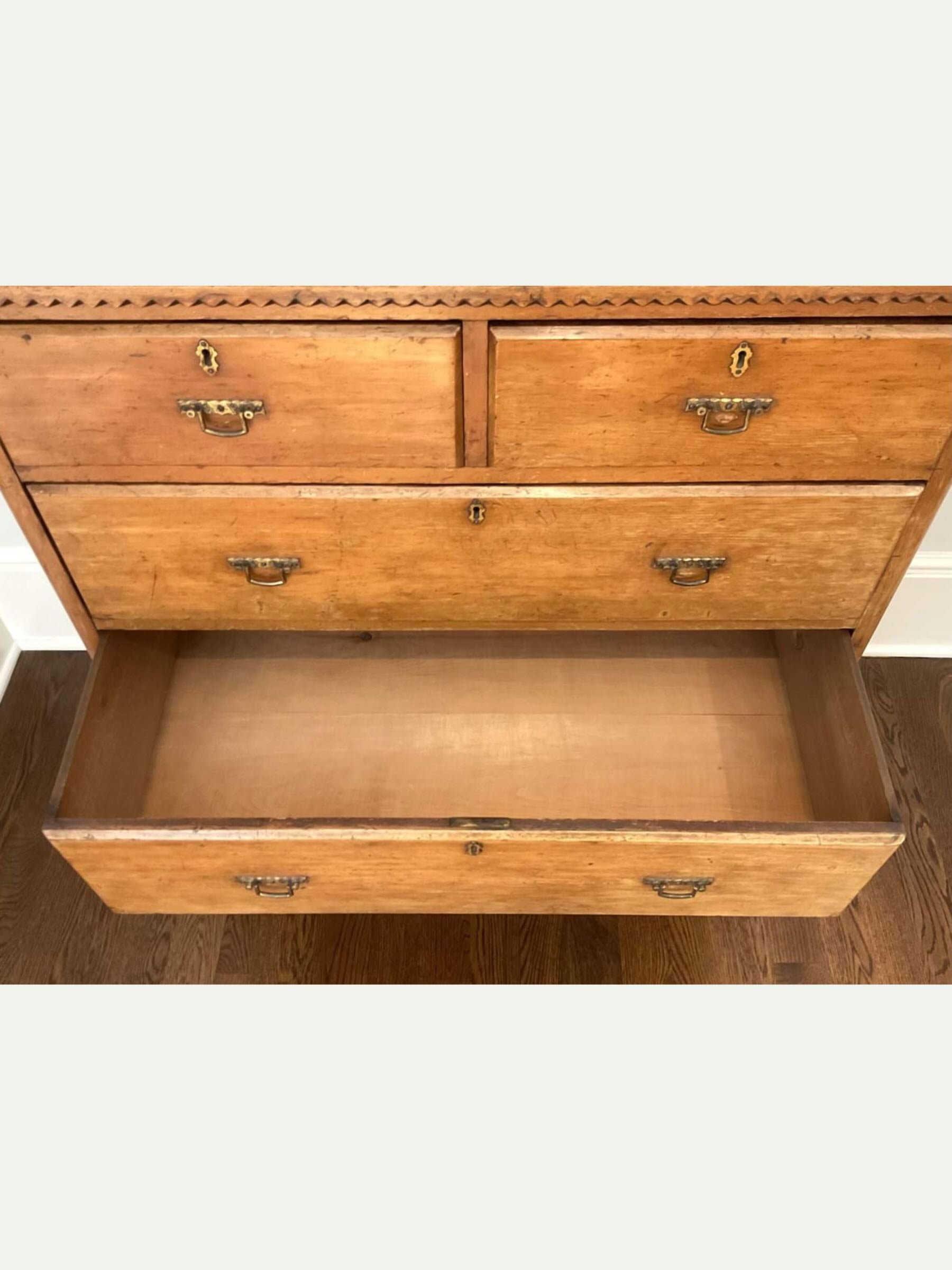 An antique wooden chest of drawers with three drawers, one pulled open, showing its interior. The drawers feature brass handles and a decorative geometric border along the top front, set against a plain light background.