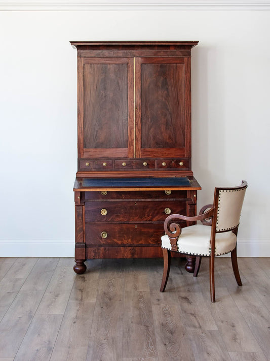 A vintage dark wood writing desk and cabinet set with an upholstered cream chair, set against a plain white wall and light wood floor.