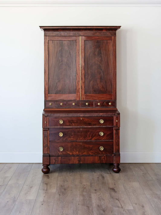 A tall antique wooden hutch or cabinet with a two-door upper section, a drawer section with brass knobs, and turned wooden feet, photographed against a plain white wall and light wood floor.