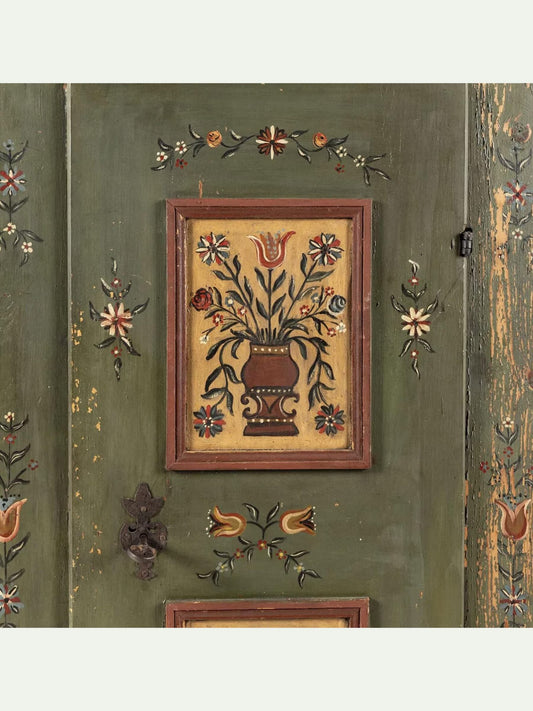 A square framed folk art panel depicting a floral bouquet in a brown vase, mounted on a distressed green wooden door with hand-painted floral motifs.