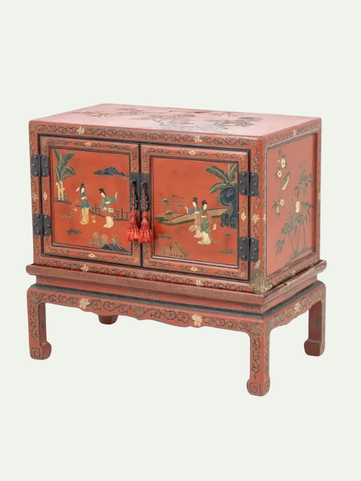 A small red wooden cabinet with two doors, decorated with hand-painted figures and scenes in black, green, and beige tones, standing on a matching carved base against a light gray background.
