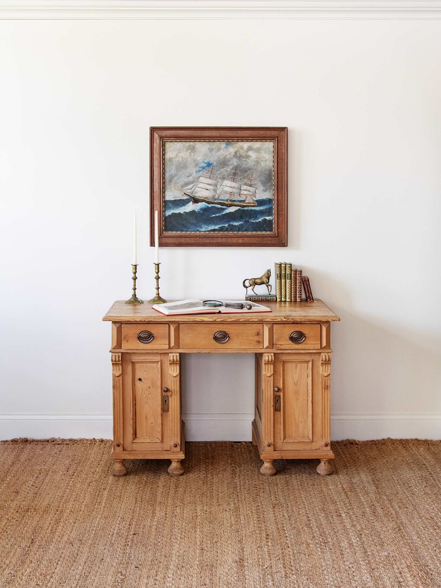 A rustic light brown wooden writing desk with three drawers and two cabinet doors, styled with a framed sailing ship painting on the wall above it, set on a woven jute rug against a white wall.