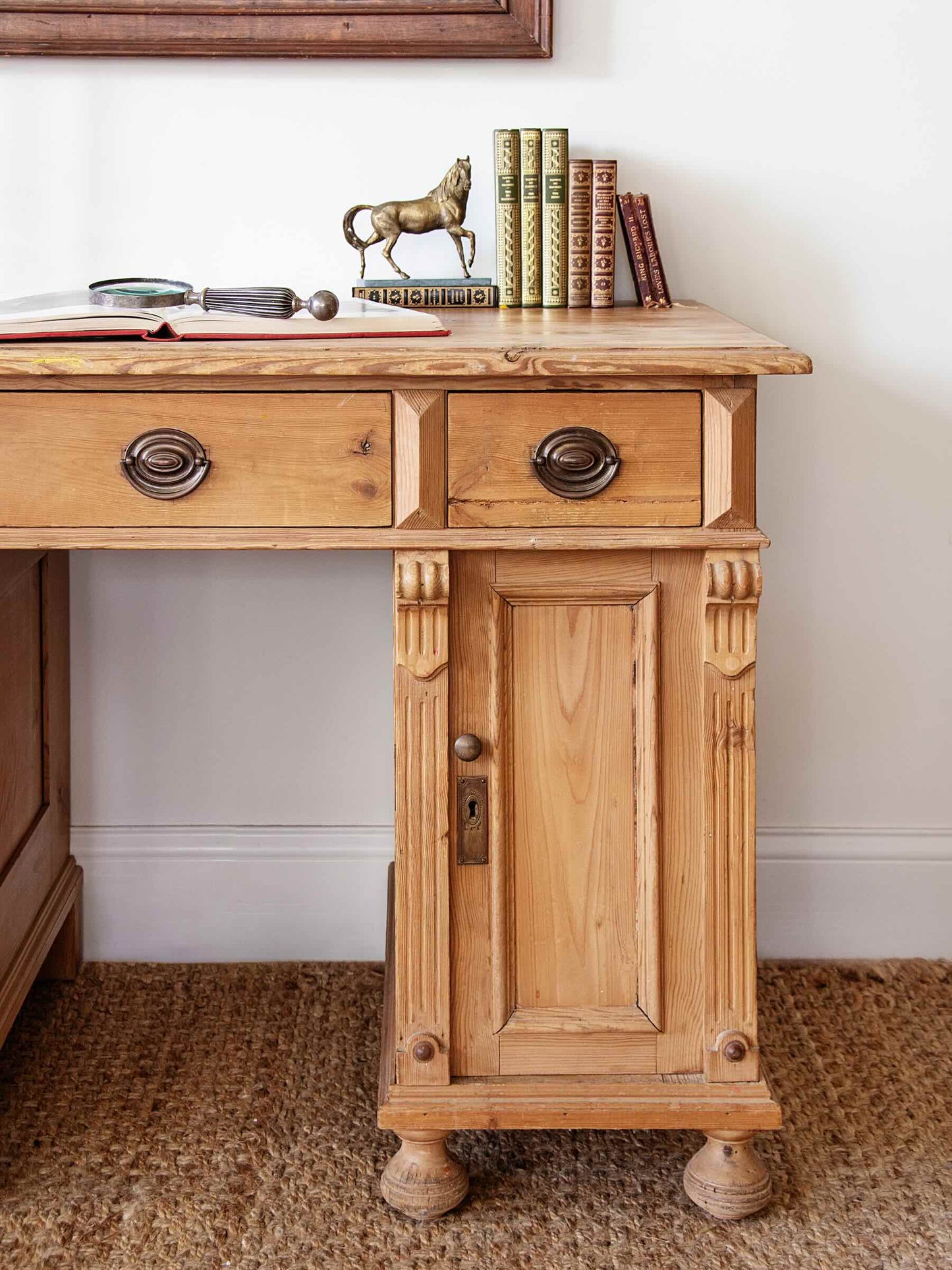 A rustic light oak wood writing desk with two drawers and a single cabinet door, styled with books and a small horse figurine on top against a white wall.