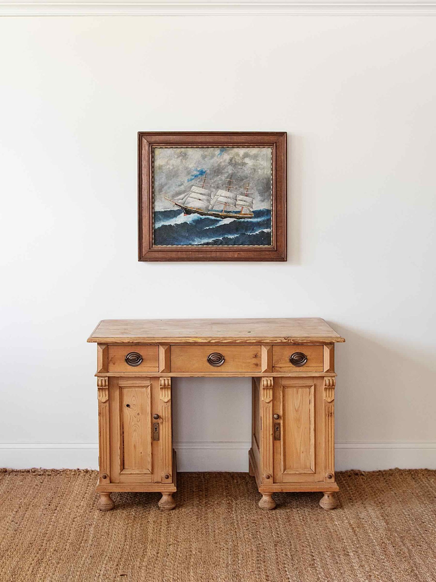 A rustic light brown wooden writing desk with three drawers and two cabinet doors, styled on a woven jute rug against a white wall with a framed painting of a sailing ship above it.