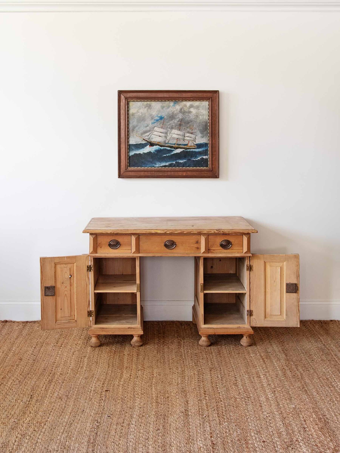 A rustic light oak wood writing desk with three drawers and open cabinet doors, styled on a woven jute rug against a white wall with a framed painting above.