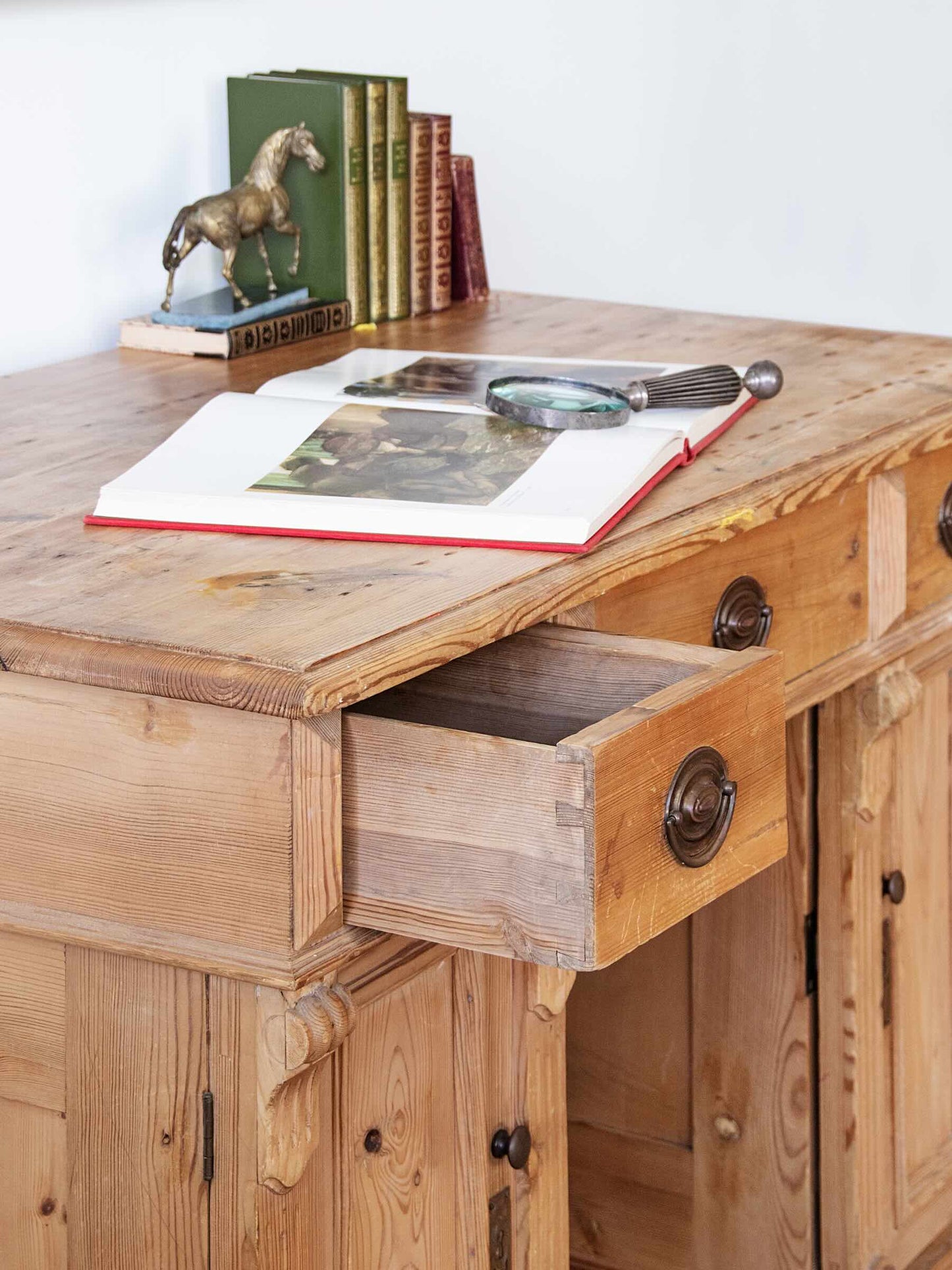 A rustic pine writing desk with a single open drawer, styled with books, a small horse figurine, and an open book with a magnifying glass on top, set against a plain white wall.