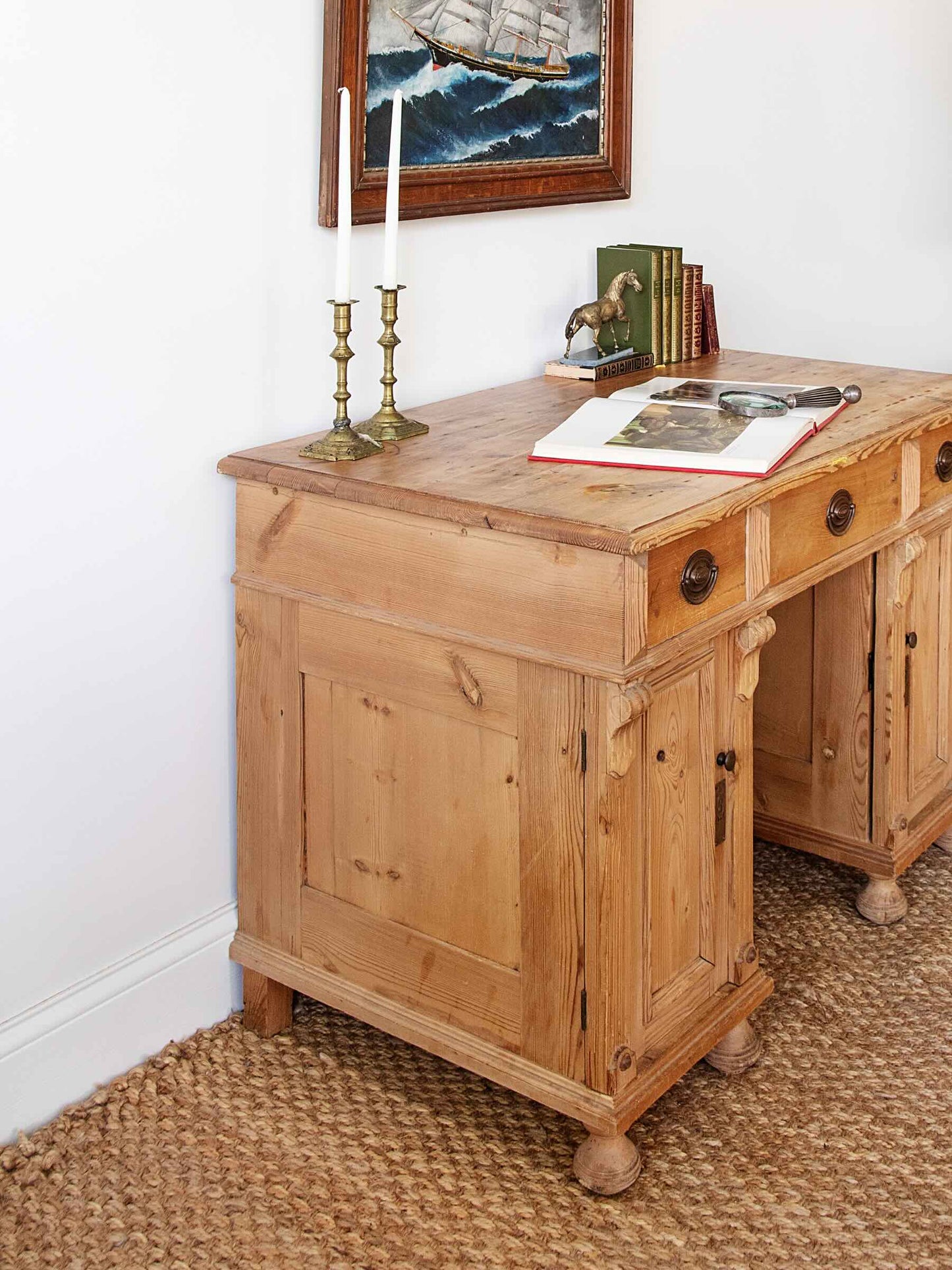 A rustic light brown wooden writing desk with drawers and turned legs, styled with brass candlesticks, books, and an open book on a woven jute rug against a white wall.