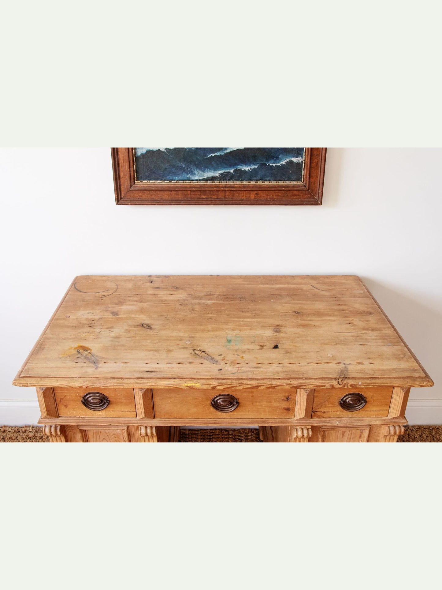 A rustic light brown wooden writing desk with three drawers and dark metal knobs, set against a white wall with a framed landscape painting above.