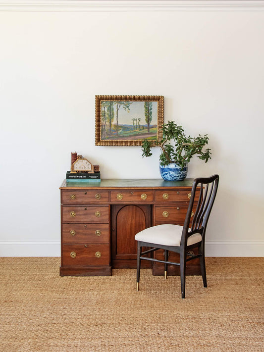 Wooden desk with chair, framed picture, and plants against a white wall.