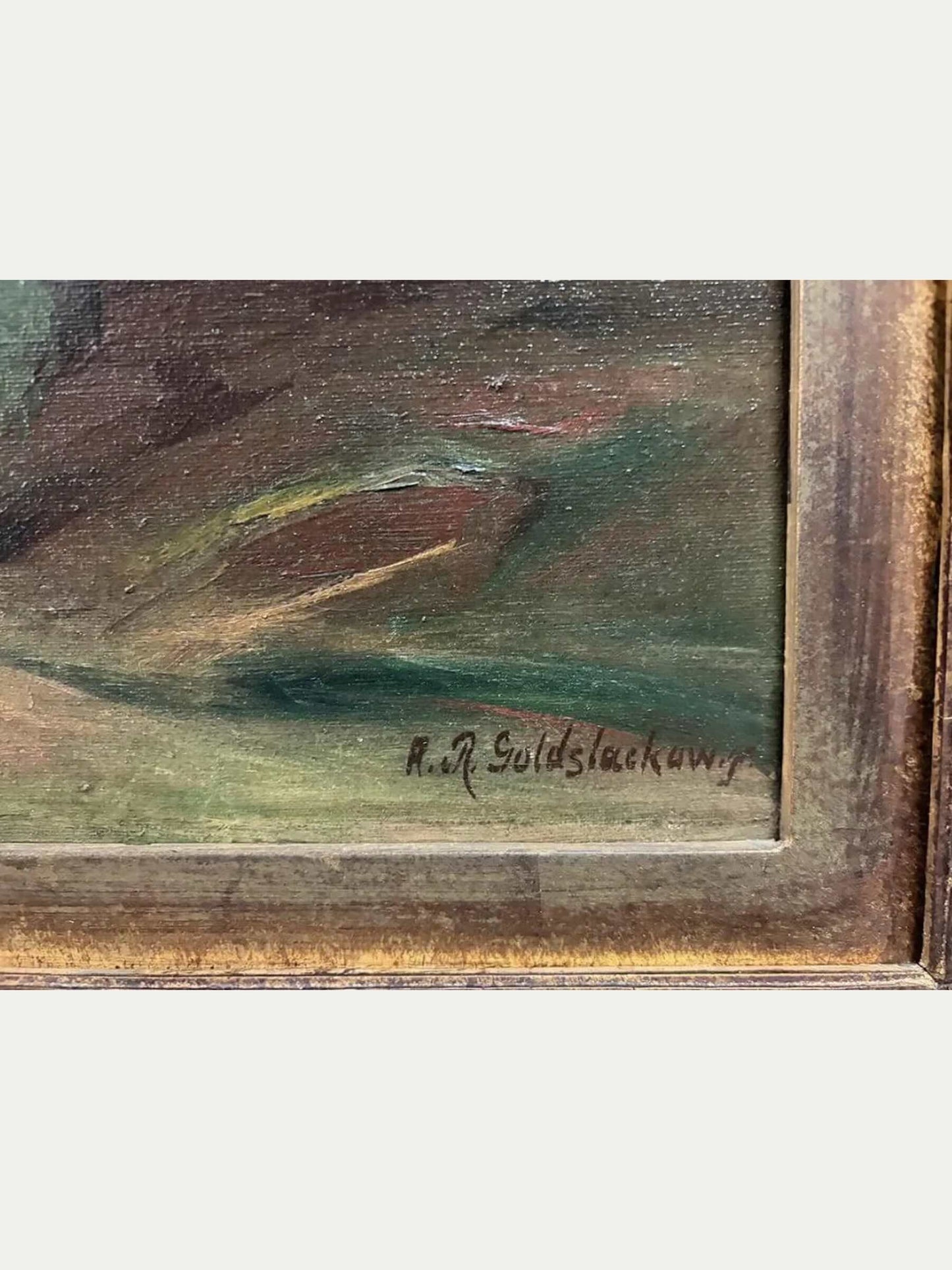 Close-up of a framed painting showing an abstract landscape or landscape-like scene with brushstrokes in brown, green, and muted gold tones, plus a visible artist signature near the lower right of the painted area.