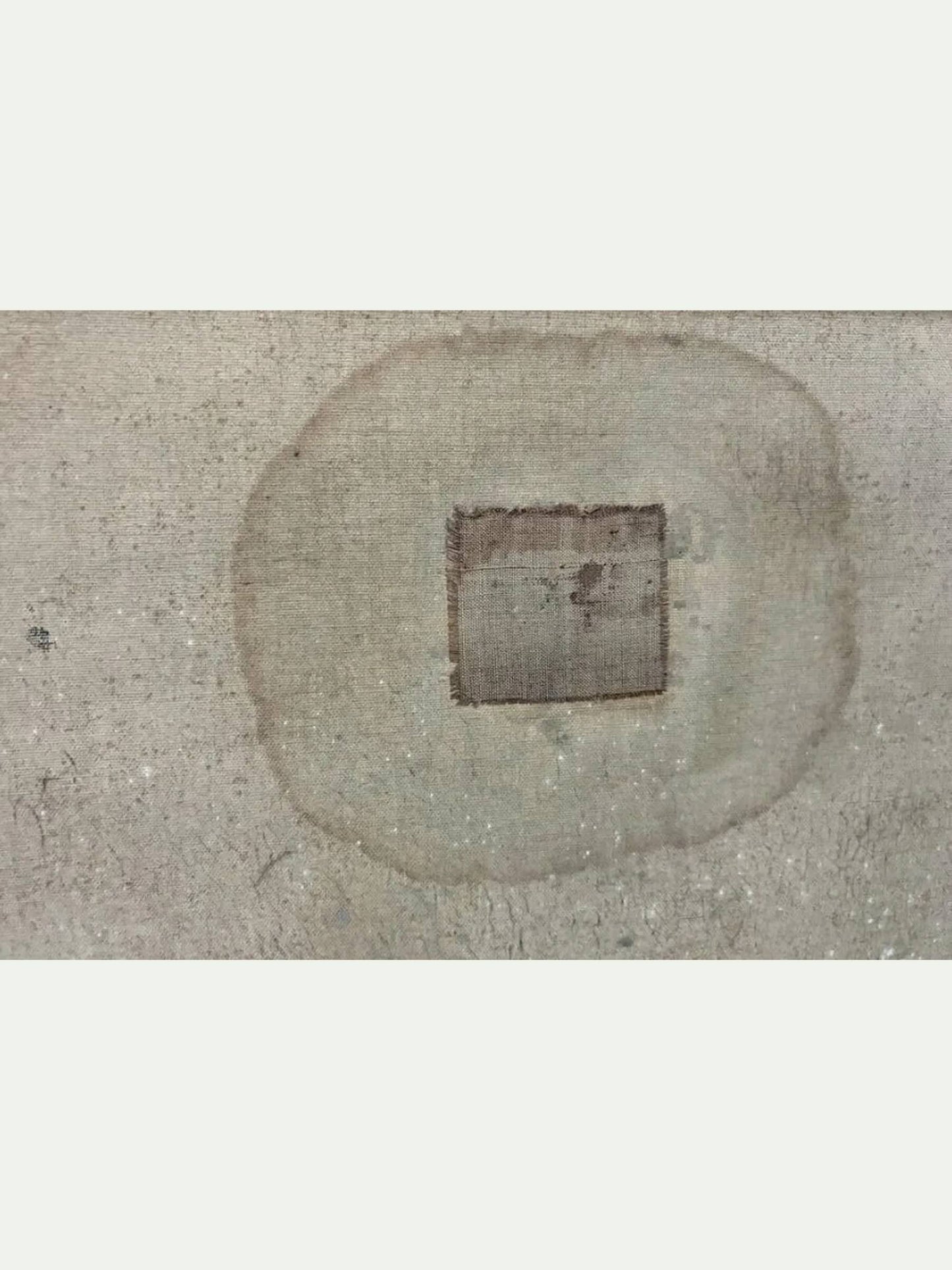A square metallic coin with a central square hole, characteristic of a Chinese Han dynasty silver ingot, placed in the center of a circular light brown stain on a textured gray concrete surface.